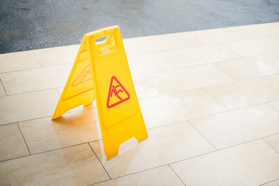 A yellow wet floor caution sign with a black and red warning icon is placed on a beige tiled floor in an indoor space. The tiles are clean, smooth, and glossy, reflecting light from above. The sign indicates potential slipping hazards and is positioned in front of a wall or entryway area. The lighting is bright and evenly distributed, highlighting the surface's cleanliness. This setup is typical for commercial or residential environments during or after cleaning or maintenance activities, and it reflects the professional cleaning services offered by Carpet Cleaning Barnes, supporting thorough surface cleaning and sanitisation for end of tenancy cleans in Barnes.