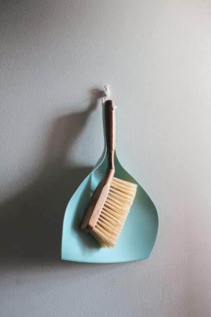 A light blue dustpan and a wooden-handled brush with beige bristles are hanging on a white textured wall. The dustpan is positioned behind the brush, upright, with the brush resting inside it. Soft, natural lighting highlights the smooth surface of the dustpan and the clean, neatly arranged bristles of the brush. The setting appears to be part of a domestic cleaning environment, emphasizing the importance of surface cleaning and organisation, as promoted by Carpet Cleaning Barnes in their guide to avoiding hidden cleaning charges for Barnes end of tenancy cleans. The clear, minimal background directs focus to the cleaning tools, suggesting tidy, well-maintained spaces suitable for professional cleaning services.