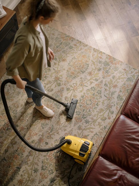 A person using a yellow vacuum cleaner to clean a patterned area rug in a living room setting. The rug features intricate floral motifs in muted tones, set on a wooden floor. The individual, dressed in casual clothing including a beige blazer and white sneakers, stands next to a red leather sofa on the right. Natural light illuminates the space, highlighting the clean and well-maintained surface of the rug. This scene depicts a professional domestic cleaning process, emphasizing surface cleaning and deep cleaning methods associated with carpet and rug maintenance, as offered by Carpet Cleaning Barnes. The image reflects hygiene standards and thorough cleaning practices typical of services explained in the Barnes carpet cleaning prices guide.