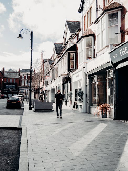 A commercial street scene showing a wide paved pedestrian walkway in front of a row of retail shops with large glass windows and white facades. The buildings feature traditional pitched roofs, some wi