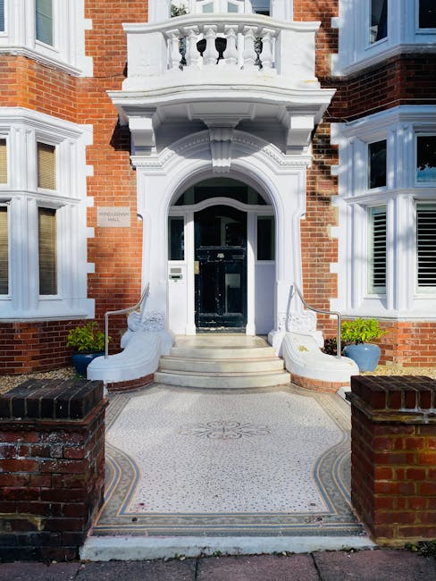 The entrance of a residential property featuring a black front door set within an ornate white stone archway. The building has red brick exterior walls with large white-framed sash windows on either side of the doorway. A curved stone staircase with white balustrades leads up to the front door, flanked by two large blue planters containing green plants. The paved area in front of the steps has intricate mosaic details, and the scene is illuminated by natural daylight, highlighting the clean and well-maintained appearance of the exterior. Carpet Cleaning Barnes offers surface cleaning and deep cleaning services for both residential and commercial spaces, ensuring hygiene and upkeep of domestic and professional settings.