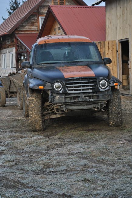 The image shows a front view of a black and orange off-road vehicle parked on a muddy ground in a rural setting. The vehicle has dirt and mud splashes on its tires and lower body, indicating recent off-road activity. In the background, there are wooden buildings with red metal roofs and unfinished walls, typical of a countryside farm or workshop area. The scene is outdoors with natural lighting, emphasizing the vehicle's use in rugged conditions. This visual does not directly relate to surface cleaning or sanitisation services but depicts a workspace environment in need of thorough cleaning and maintenance, which could be offered by Carpet Cleaning Barnes as part of their domestic cleaning and deep cleaning services near Barnes Bridge and Lonsdale Road.