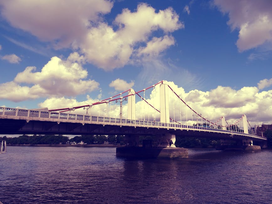 A large suspension bridge spanning a wide river, featuring white towers and red suspension cables, with a blue sky dotted with fluffy white clouds in the background. The bridge's roadway appears clean and well-maintained, with no visible debris or dirt, and the water below is calm, reflecting the sky and structure. The image showcases a bright, sunny day with natural lighting illuminating the scene, suitable for surface cleaning or exterior maintenance discussions related to the services offered by Carpet Cleaning Barnes near Barnes Bridge and Lonsdale Road.
